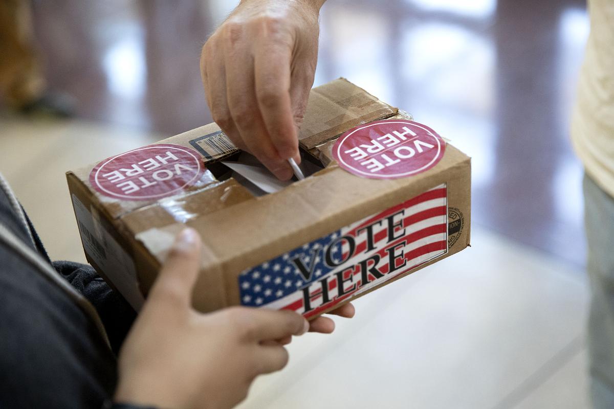 A hand drops a ballot into a brown box with the word "vote here" written on it. 