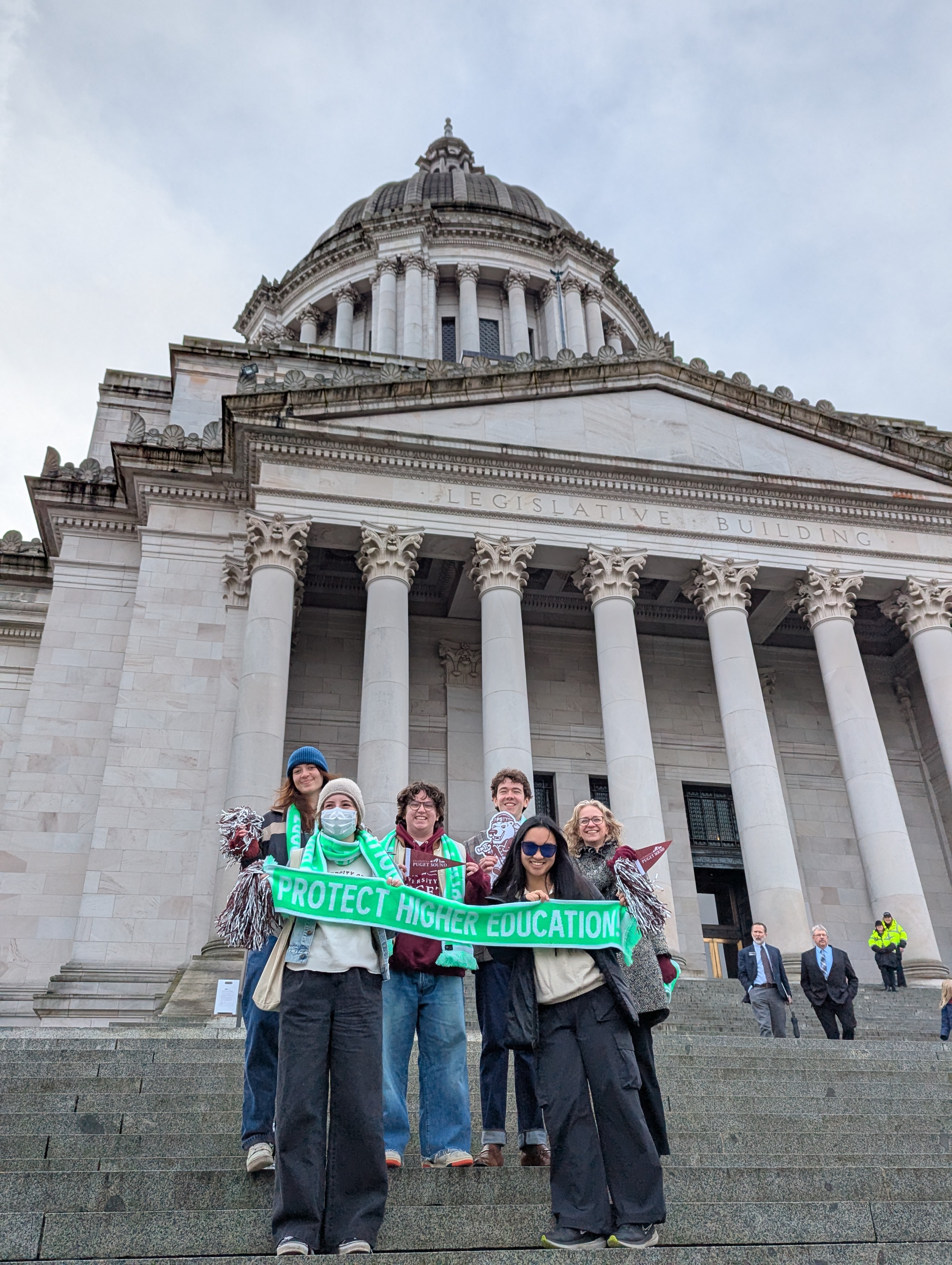 Five University of Puget Sound students and Prof. Robin Jacobson stand on the steps of the Washington State Capitol holding a green scarf with the words "Protect Higher Education" on it. 