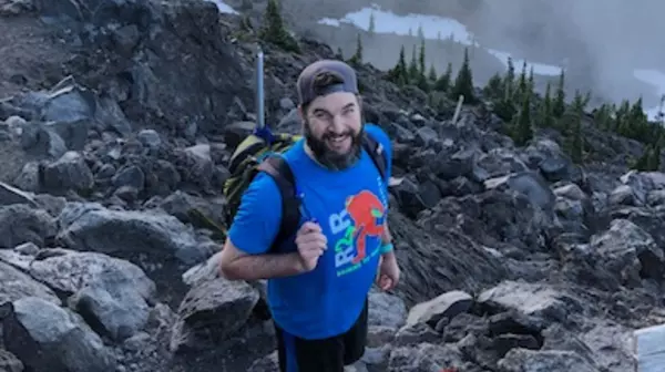 Dr. Bryan Thines with a backpack on, standing on a rocky slope with sparse pine trees, some snow and fog behind him. 