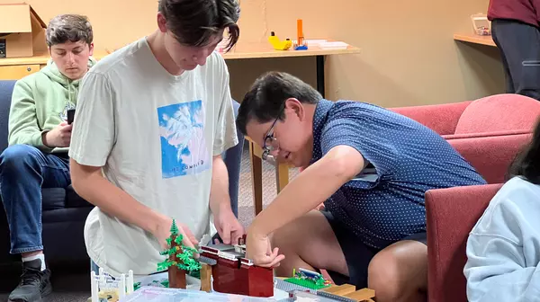 Students facing down at a table with a Lego structure, reach out to continue building.