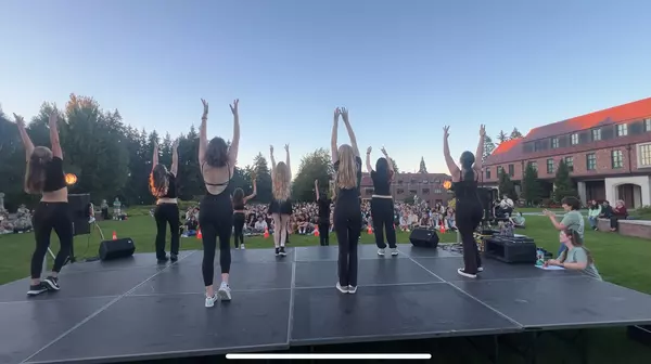 Students dressed in black clothing stand with arms raised above them on a stage in a grassy field with people sitting on the grass in front of them.