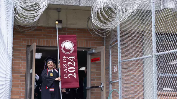 An incarcerated woman wearing a graduation gown and holding a Class of 2024 banner leaves a building between fences toped with razor wire.