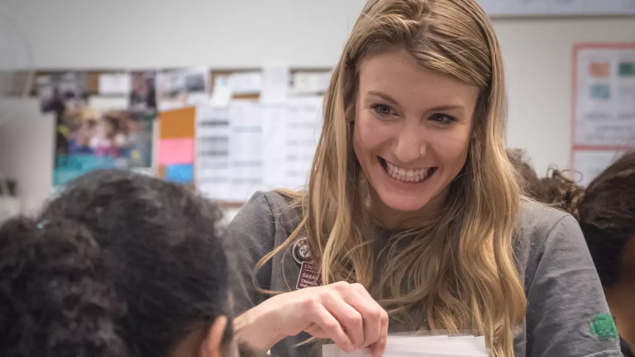 A person smiling and handing out papers to kids in a classroom