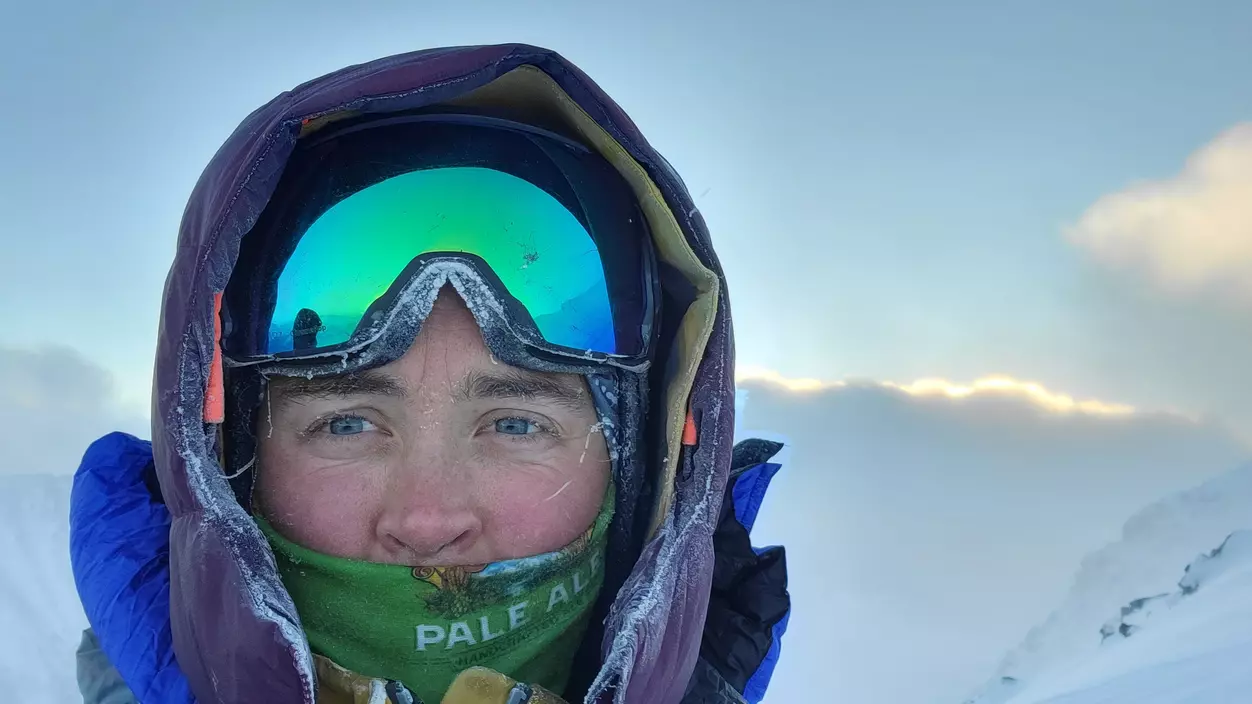 A woman in climbing gear, the lower half of her face covered and her reflective goggles pulled up onto her forehead.