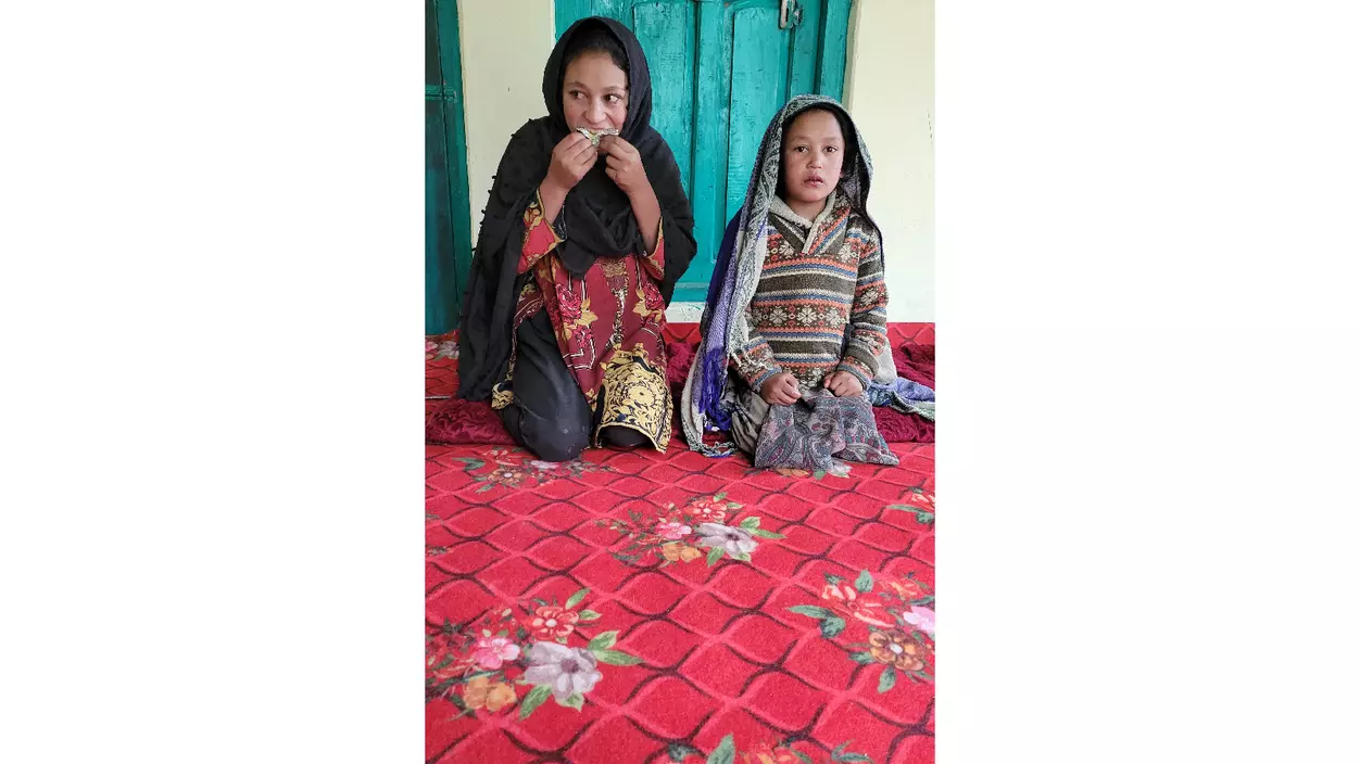 Two young girls in boldly patterned dresses and head scarves kneel on a red rug. One appears to be eating something.