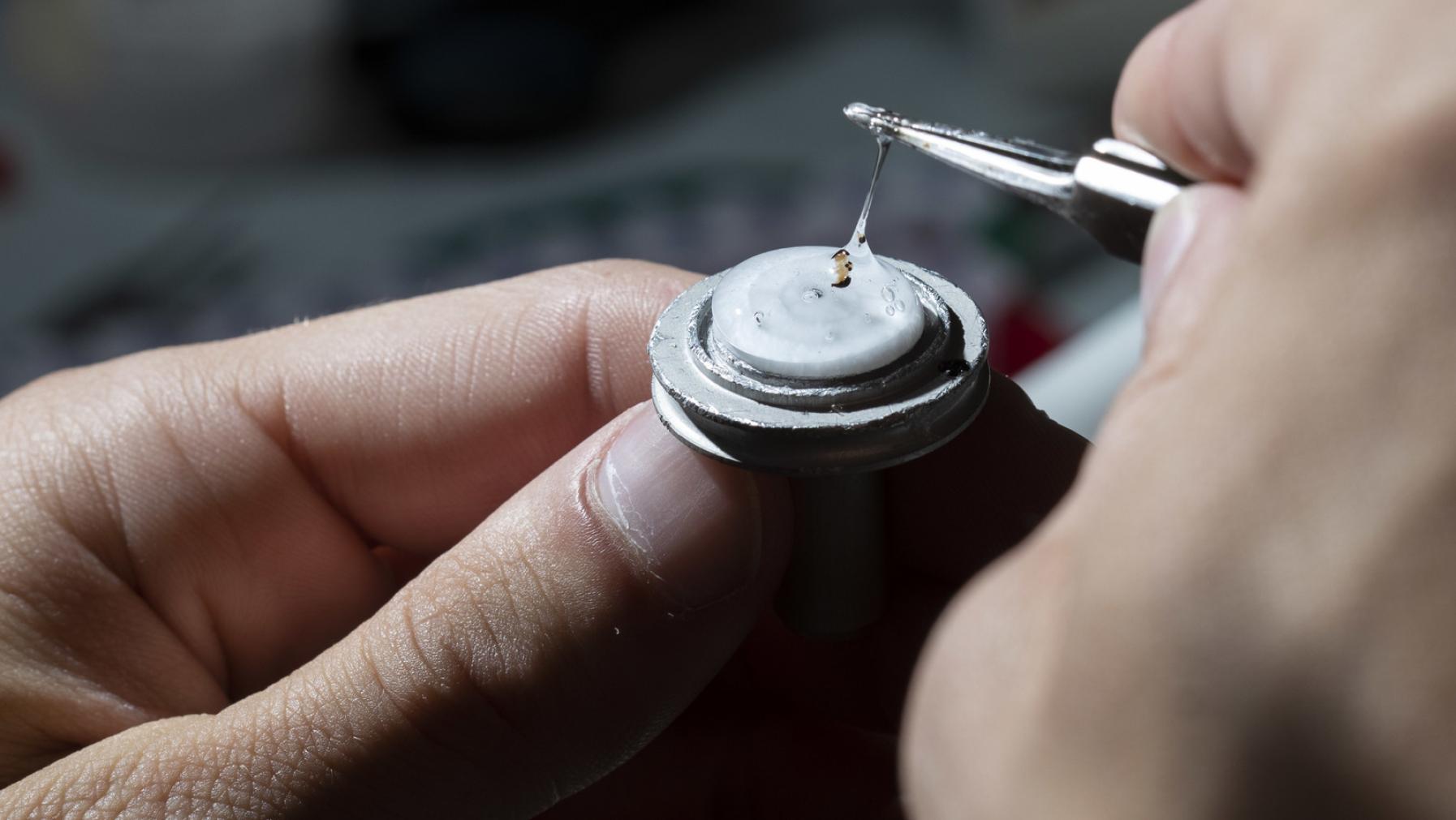 Closeup of a hand holding a circular disc with a thick, clear liquid, while a second hand carefully places a tiny specimen into the liquid with delicate tweezers