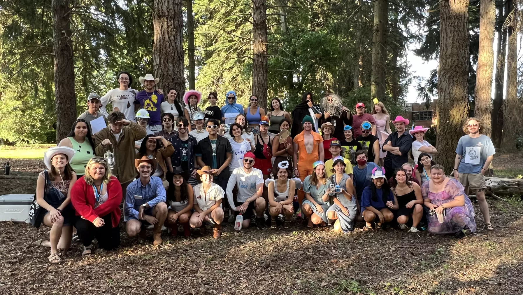 A large group of students in various costumes pose together in a forest. Some stand on a long fallen tree truck.