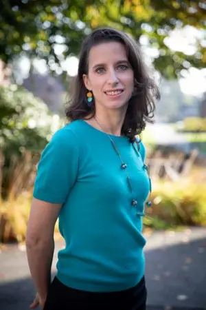 A photo of Professor Sara Protasi taken outside. Sara is standing with trees in the background, wearing a blue shirt, funky necklace, and colorful earrings. There is no text in the image. 