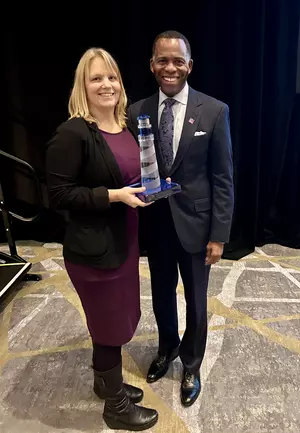 A blonde woman in a maroon dress and black blazer stands next to a man in a dark suit holding a lighthouse trophy. 