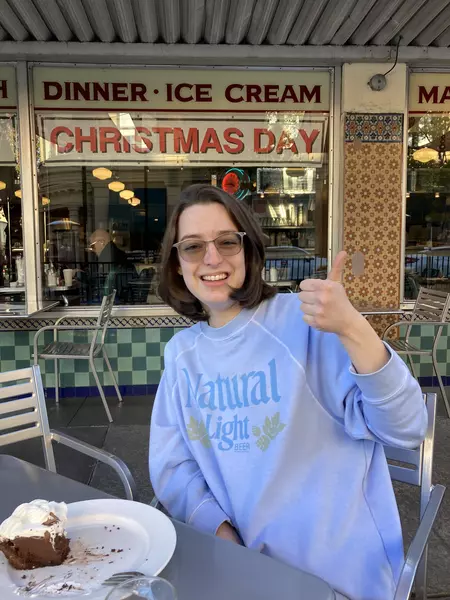 Photo of Svetlana sitting in front of a diner with a slice of cake in front of her. She is giving the camera a thumps-up