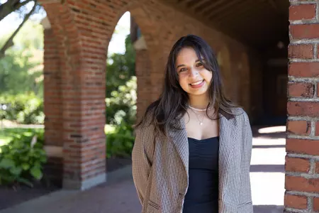 Maya Gomez faces the camera with her hair tilted. She is standing under red brick arches and is wearing a blazer. 