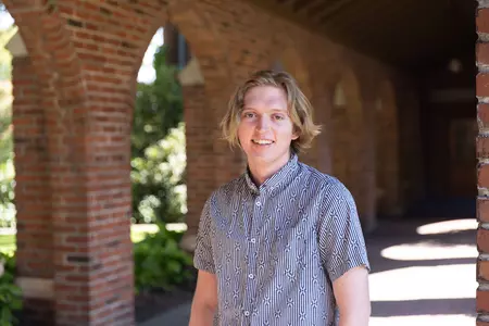 Oliver Wright stands under red brick arches facing the camera. He has blonde hair and is wearing a blue short-sleeve shirt. 